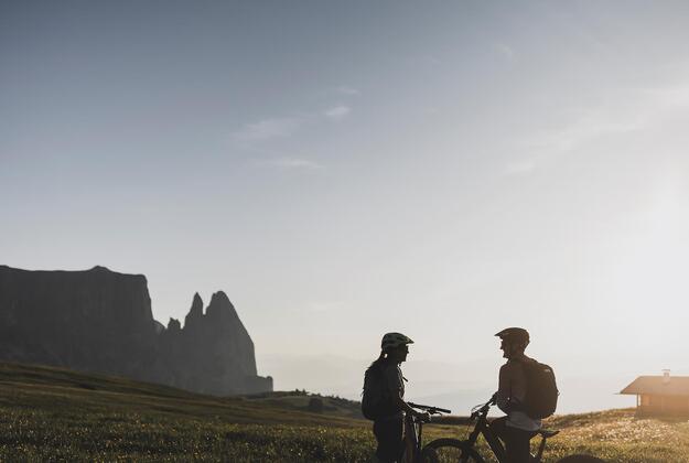 Coppia in e-bike al tramonto sull'Alpe di Siusi con la sagoma dello Sciliar